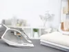 A steam iron and folded clothes on an ironing board, with a blurred background of a room. Produktbild-Vorschau