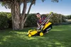 A woman is using a yellow and black Kärcher lawnmower on a green lawn in front of a large tree and bushes. Produktbild-Vorschau