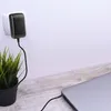 A laptop is charging via a power adapter plugged into a wall socket, with a potted plant nearby. Voir les images du produit