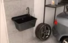 A black sink with a faucet is mounted on a tiled wall in a garage or workshop. A car tire and part of a car are visible next to it, along with a shelf with bottles. Náhled obrázku produktu