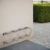 A metal bicycle rack is mounted on a textured wall next to a neatly trimmed green hedge and a paved walkway. Náhled obrázku produktu