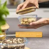 A person is holding a glass food container filled with peanuts, with a bamboo lid being placed on top. Other containers with food are visible in the foreground. Produktbild-Vorschau