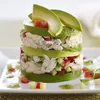 A layered dessert or appetizer made with avocado, crab salad, and diced tomatoes and cucumbers, presented on a white plate with a drizzle of green sauce. Produktbild-Vorschau