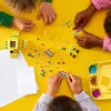 Children's hands assembling colorful LEGO DOTS pieces on a yellow surface. Podgląd zdjęcia produktu