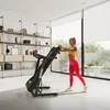 A woman is exercising on a treadmill in a modern, well-lit room with large windows and shelves filled with decor. Produktbild-Vorschau
