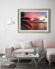 A framed picture of a sunset over the ocean with palm trees in the foreground, displayed above a pink sofa in a living room. Produktbild-Vorschau