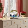 A festive Christmas scene displayed on a dresser, featuring a miniature animated village with lights and figures, surrounded by decorative elements like reindeer and poinsettias. Náhled obrázku produktu