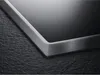 A close-up of the corner of a black glass ceramic hob with a brushed metal frame, resting on a dark, textured surface. Náhled obrázku produktu