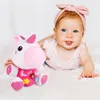 A baby girl is lying on a white surface next to a plush unicorn toy. Anteprima dell’immagine prodotto