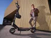 A man leans against a building with an electric scooter next to him. Produktbild-Vorschau