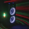 A tall, black speaker with two illuminated woofers is shown in a dark room with red and green light effects projected onto the wall and floor. Voir les images du produit