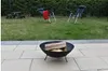 A fire pit with logs sits on a patio in front of a grassy lawn with chairs and a picnic table in the background. Produktbild-Vorschau