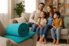 A family is sitting on a couch in a living room, with a rolled-up turquoise blanket in the foreground on a chair. Produktbild-Vorschau