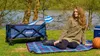 A woman sits on a picnic blanket next to a blue foldable cart by a lake. Produktbild-Vorschau