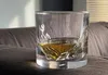 A glass of whiskey sits on a dark fabric surface, with a blurred background of a couch and window blinds. Produktbild-Vorschau