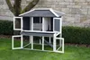 A wooden rabbit hutch is set up on a green lawn in front of a stone wall and a tree. Voir les images du produit