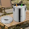 A stainless steel insulated food carrier filled with ice and bottles of drinks, placed on a wooden table outdoors, with camping chairs in the background. Produktbild-Vorschau