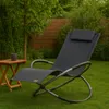 A dark gray rocking lounge chair sits on a green lawn next to a small wooden table with a mug on it. A wooden fence and trees are in the background. Produktbild-Vorschau