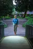 A woman walks down a path in a park at dusk, holding a flashlight. Náhled obrázku produktu