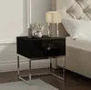 A black bedside table with a silver frame, a drawer with a crystal knob, a lamp, and a vase with flowers. The bedside table is next to a bed with a tufted headboard. Produktbild-Vorschau