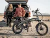 A couple sits on the tailgate of a vehicle with an electric bike parked next to them in a desert-like environment. Produktbild-Vorschau
