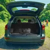 The open trunk of a car with a black bag inside, parked in a grassy field with two people in the background. Produktbild-Vorschau