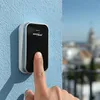 A finger presses a wireless doorbell mounted on a blue wall, with a blurred cityscape in the background. Náhľad obrázku produktu