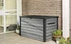 A storage box is placed on a patio next to a brick wall and a glass door. Produktbild-Vorschau