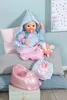 A baby doll with accessories, including a hooded robe, a pacifier, a small toy, a potty, and a pair of bloomers, is arranged on pillows. Produktbild-Vorschau