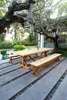 A wooden dining set with benches is placed on a patio next to a swimming pool, under a large tree with hanging lanterns. Produktbild-Vorschau