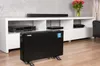 A black electric heater is placed on a wooden floor in front of a white entertainment center with electronics and a vase of flowers. Podgląd zdjęcia produktu