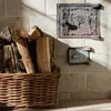 A basket of firewood next to a tiled wall with a decorative metal plate. Produktbild-Vorschau