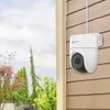 A white EZVIZ security camera mounted on a brown wooden wall, with a garden and sky visible through a window on the left. Produktbild-Vorschau