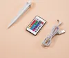 A white ground stake, a remote control with colorful buttons, and a gray USB charging cable are laid out on a light beige background. Produktbild-Vorschau