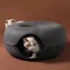 Two cats are in and around a round, dark gray cat bed. One cat is peeking out from the opening of the bed, and another cat is visible in the background. Produktbild-Vorschau
