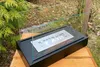 A black tabletop bio-ethanol fireplace with a glass wind guard sits on a wooden surface outdoors, surrounded by greenery and fallen leaves. Produktbild-Vorschau