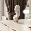 A white bust sculpture with a gold base sits on a white vanity table, with brown curtains and illuminated vanity lights in the background. Produktbild-Vorschau