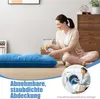 A woman is kneeling on the floor next to a blue floor mattress, pointing at it. In the background, there is a washing machine with a person loading clothes. Produktbild-Vorschau