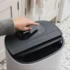 A person's hand is shown pressing a button on the control panel of a Meaco dehumidifier, which is placed in a domestic setting with cabinets and patterned tiles visible in the background. Náhled obrázku produktu