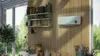 A wall-mounted heater is shown in a sunlit room with shelves of decorative items and a large window overlooking a garden. Produktbild-Vorschau