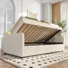 A beige sofa bed with its storage compartment open, revealing the slatted base. The bed is in a well-lit room with a large window and curtains in the background. Produktbild-Vorschau