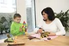 A woman and a child are sitting at a table, engaged in a craft activity. The child is holding a tower of colorful blocks, while the woman is using a glue gun to decorate a black and yellow striped object. Produktbild-Vorschau