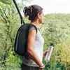 A woman wearing a backpack holds a water bottle while looking out at a natural landscape. Produktbild-Vorschau