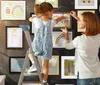 A mother and daughter are hanging children's drawings in frames on a wall. Produktbild-Vorschau