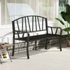 A black metal garden bench sits on a light-colored patio in front of a building with large windows and a stone wall. Voir les images du produit