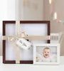 Two photo frames, one larger and one smaller, are displayed on a surface. The larger frame is dark brown and wrapped with a beige ribbon and a 'For You' gift tag. The smaller frame is white and contains a picture of a smiling baby. Produktbild-Vorschau