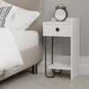 A white bedside table with a drawer and a shelf, next to a bed. An alarm clock sits on top of the table.