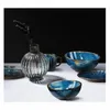 A collection of blue ceramic tableware and a glass vase with dried flowers on a white surface against a black background. Produktbild-Vorschau