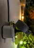 A smart plug with a protective cover is plugged into an outdoor outlet, surrounded by green foliage. Produktbild-Vorschau