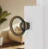 A close-up of a bronze ring pull on a white piece of furniture, with a blurred background of a room. Anteprima dell’immagine prodotto